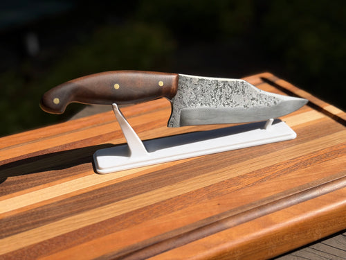 A chef's knife with a textured Damascus steel blade and a brown wooden handle, displayed on a white stand over a striped wooden cutting board.