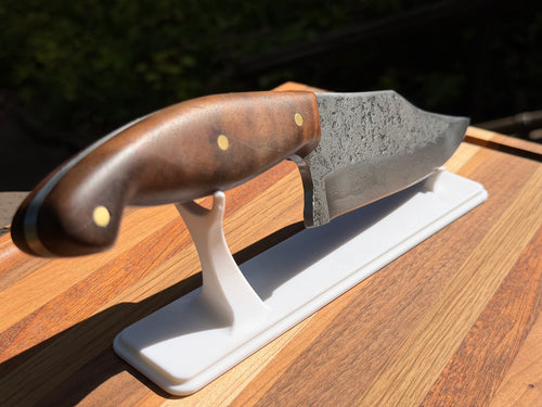 A large fixed-blade knife with a dark textured steel blade and a brown wooden handle secured by brass pins, displayed on a white stand on a wooden surface.