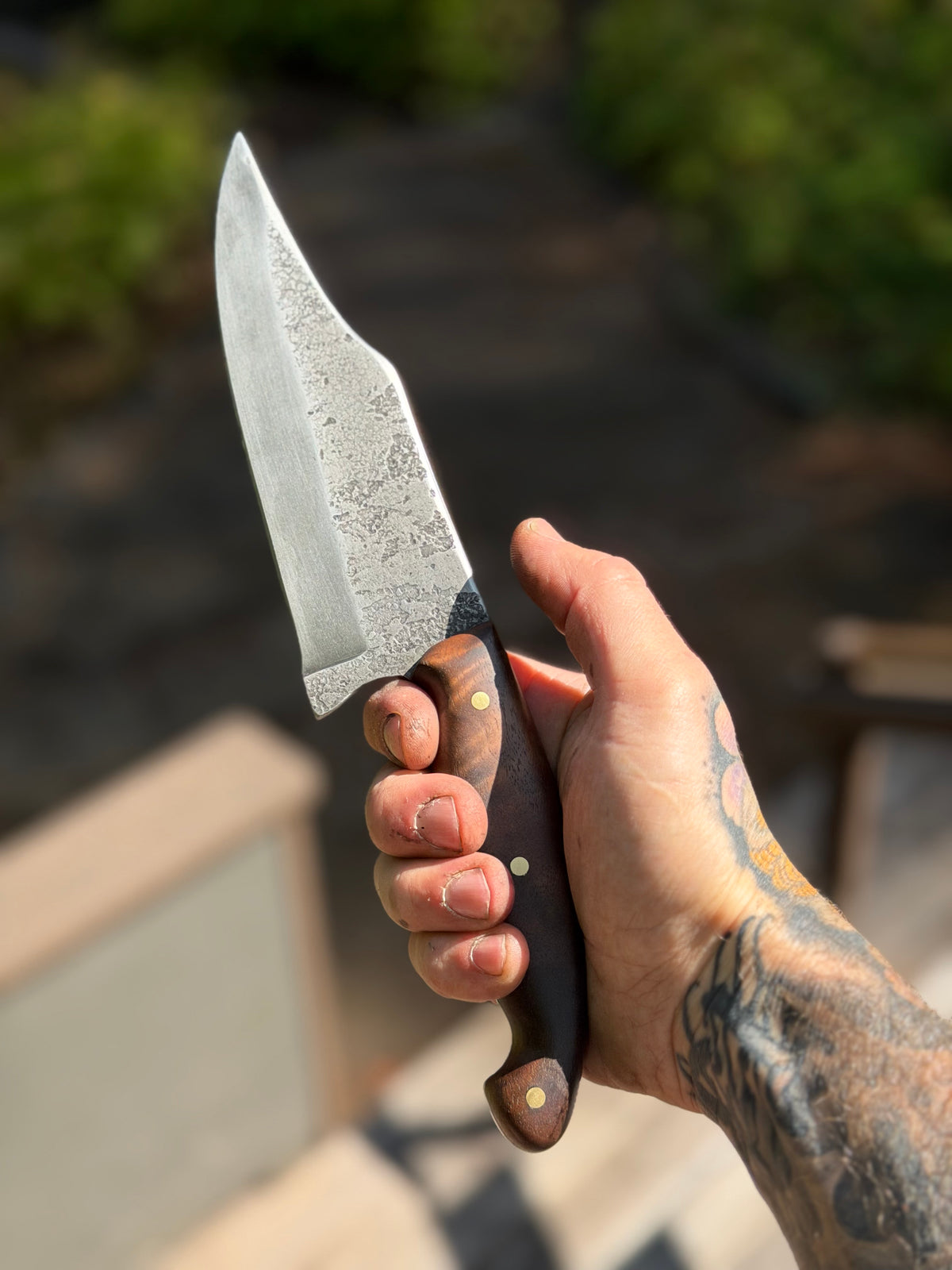 A hand with tattoos holds a fixed-blade knife with a textured, hammered steel blade and a dark brown wooden handle with brass rivets, set against a blurred outdoor background.