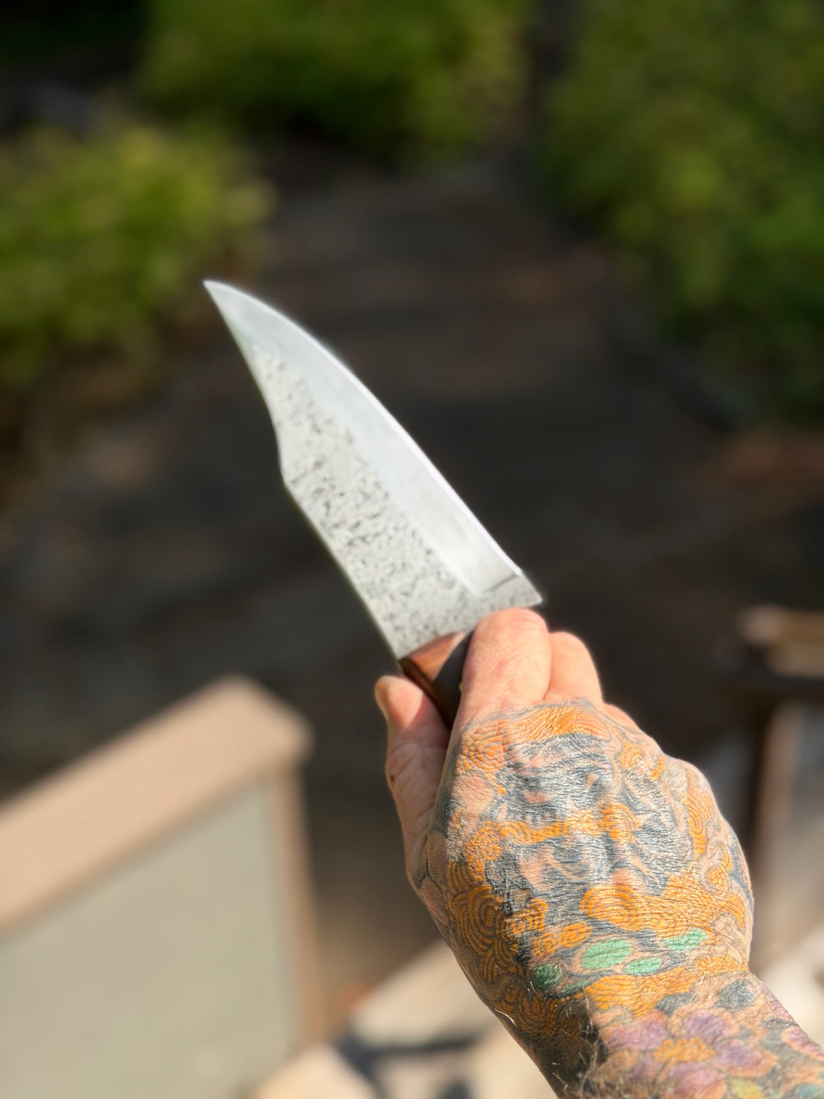 A hand with colorful tattoos holds a fixed-blade knife with a textured silver blade and a dark handle, set against a blurred outdoor background.