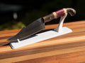 A curved chef's knife with a dark wood handle and reddish-pink grip bands, displayed on a white stand over a wooden surface.