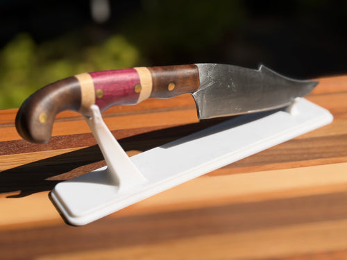 A fixed-blade knife with a dark wood handle, pink and beige inlays, and a dark steel blade, displayed on a white stand on a wooden surface.